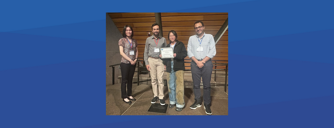 Four people standing together indoors. Two men and two women, all wearing conference badges, are posing for a photo. PhD student Zhuocheng Shang (far left) and Professor Ahmed Eldway (second from left) stand alongside two other colleagues. The person in the center is holding a certificate or award. They are all smiling.
