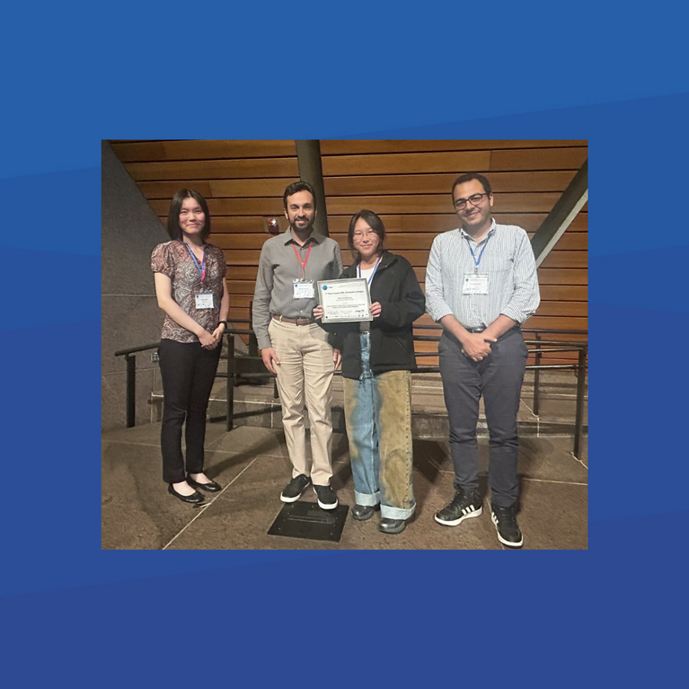 Four people standing together indoors. Two men and two women, all wearing conference badges, are posing for a photo. PhD student Zhuocheng Shang (far left) and Professor Ahmed Eldway (second from left) stand alongside two other colleagues. The person in the center is holding a certificate or award. They are all smiling.