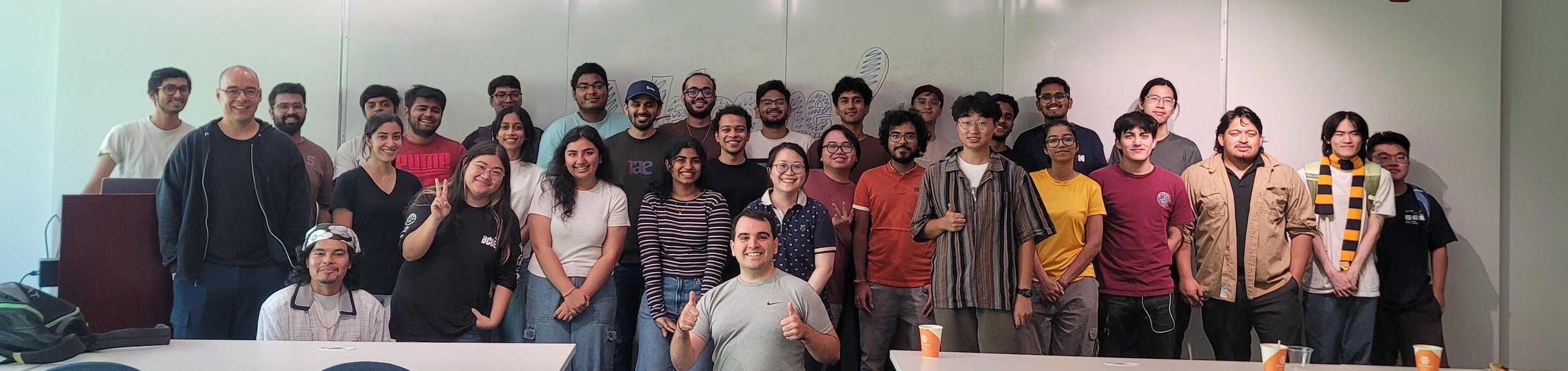 Group photo of UC Riverside Computer Science graduate student association standing together in a classroom. Around 30 people are smiling at the camera in front of a whiteboard, with tables and chairs in the foreground and pizza plates and cups on the tables.